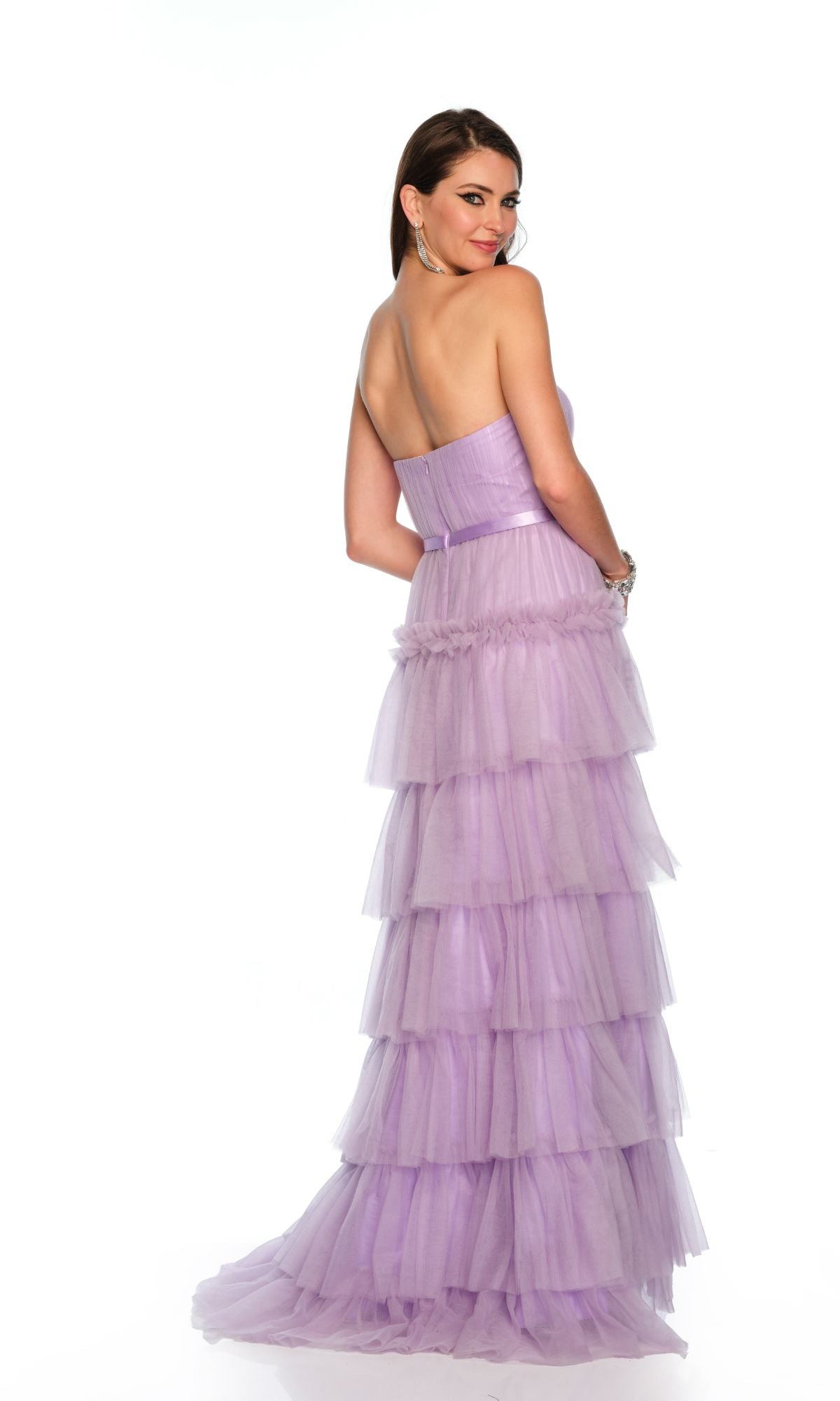 A woman in the Long Formal Dress 11579 by Dave and Johnny, featuring tiered lavender tulle and a strapless ruffle design, stands facing away and smiles at the camera against a plain white background.